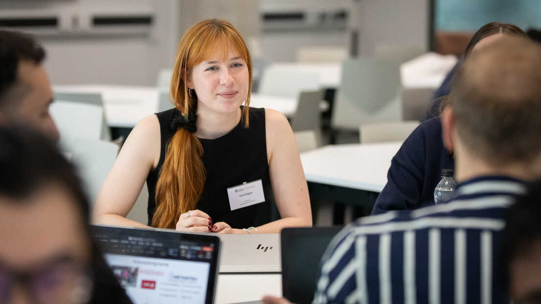 A woman with long red hair is smiling and listening in a group of people sat talking at a table with laptops