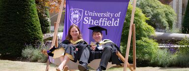 A woman and a man in graduation robes sit on an oversized deck chair