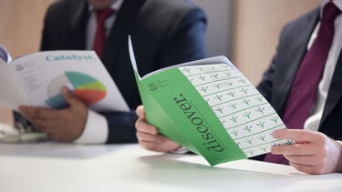 Men (can't see their faces) with open brochures. On the cover, one has a pie chart and the other has the words 'discover'.