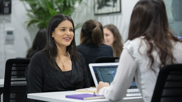 Two students talk while sitting and facing each other. A laptop is open between them. Other students can been seen in the background.