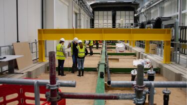 The yellow steel crossbeam and metallic structures of a large scale facility surround a group of researchers in high viz jackets and hardhats stood on sand