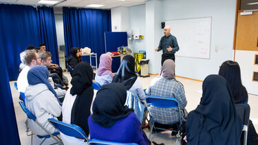 A group of students learn clinical skills from a lecturer. He is presenting to them. They are sitting down.