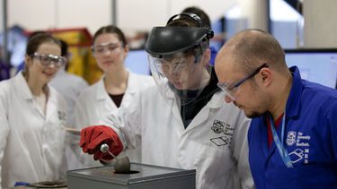 An engineering student learning in The Diamond. Two others students look on. They all have goggles and lab coats on. 