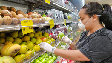 Woman wearing a mask in a supermarket and gloves. She is picking up an apple.