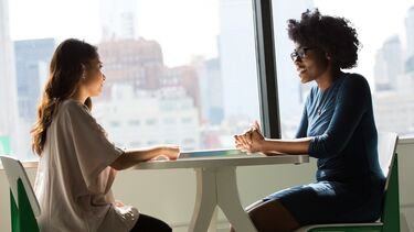 Two women having a meeting. They are sitting at a table across from each other with highrise buildings visible in the background.