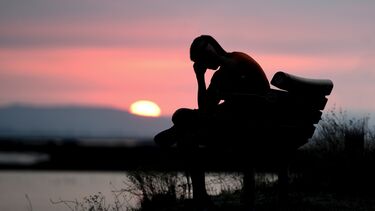 Silhouette of person sitting on a bench with their head in their hands agains a pink sky and setting sun