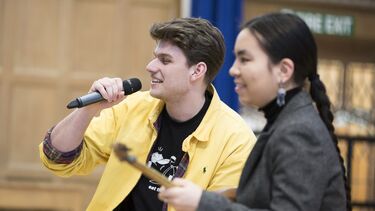 Two music students rehearsing - one is singing into a microphone, the other sites closer to the camera and plays the guitar