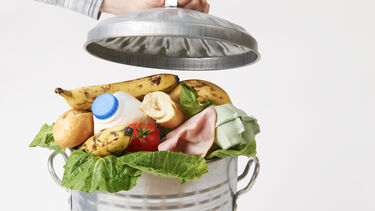 A metal rubbish bin overflowing with wasted food. A man is holding the bin lid up.
