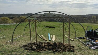 A green field with the frame of a polytunnel build on it. Under the frame are a set of wooden poles and the frame covering.