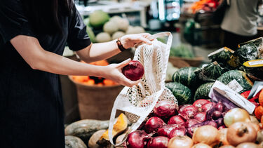Cropped shot of young Asian woman shopping for fresh organic red onions in supermarket. She is shopping with a cotton mesh eco bag and carries a variety of fruits and vegetables. Zero waste concept