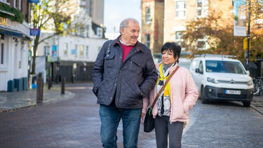 Older couple walking down the street. He is wearing a grey jacket and she is wearing a pink jacket.