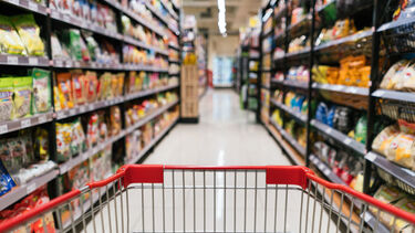 A supermarket trolley in front of full shelves of packaged foods. 