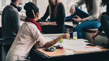 A group of people sat around a table. On the table are sheets of papers and various items of stationery.