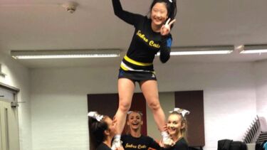 Cheryl practicing cheerleading with 3 others. Cheryl has been lifted by the others.