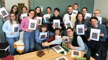 a group of about 16 students all with a script in their hands and in the middle three people cut into a cake