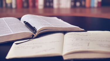 An open printed book, a pen and an open notebook on a table. In the background is a bookshelf.