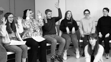 Group of students sat on chairs in a semi circle and they gesture while singing a song