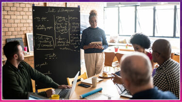 Woman stood presenting at blackboard in front of a table of adults. Informal setting