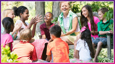 Kids surround teacher in centre of circle. Group is outside. Everyone is laughing and smiling. 