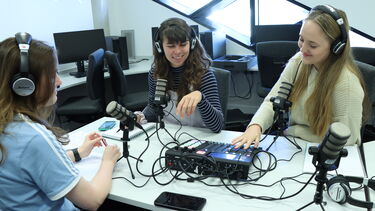 A group of students wearing headphones, sat around microphones in recording a podcast
