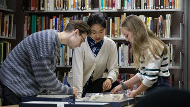 Three students looking at archive material at the Library. There is a full bookcase in the backgroud.