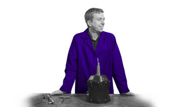 Student in a lab coat smiles and leans against a work bench. A piece of machinery is pictured in front of them, on the bench.
