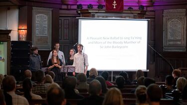 A projector screen showing "A Pleasant New Ballad to sing Ev'ning and Morn of the Bloody Murther of Sir John Barleycorn" with a band in front of an audience