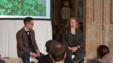 Two individuals sat in front of an audience, discussing slides as shown on a projector screen behind them