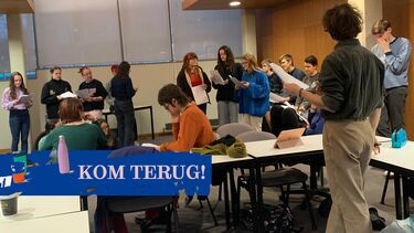 Group of about fifteen students with scripts in their hands and standing around a room with director