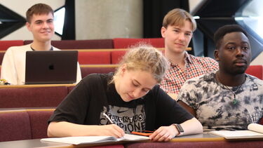 A group of Philosophy students sat in a lecture hall. One student is writing in a notebook. One student is working on a laptop.