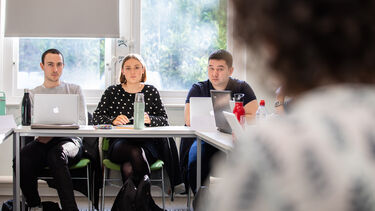 Three students in a seminar room listening to their seminar tutor