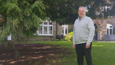 John Lavender wears a grey sweatshirt. He is stood outside an old stone building with a tree on the left.