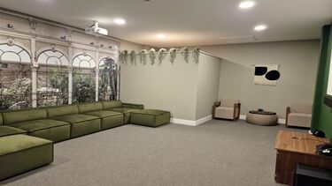 A photograph inside a room, with a large green sofa, chairs and a small table. Out of view is a projector screen. The wall is decorated to resemble the Sheffield Botanical Gardens,