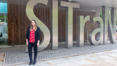 A woman with long blonde hair wears a black cardigan and red top. She stands in front of a metal set of letters spelling 'SITraN'