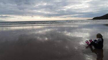 A woman wears diving flippers sat on the beach with clouds overhead.