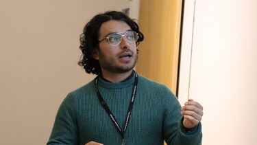 A man wearing a green jumper is standing in front of a screen lecturing