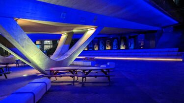The underpass of the A57 lit up in with yellow and blue lights, with stepped seating, stone benches and wooden picnic benches