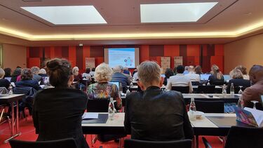 View from back of the room in a large lecture hall, red walls, people seated with backs to the camera