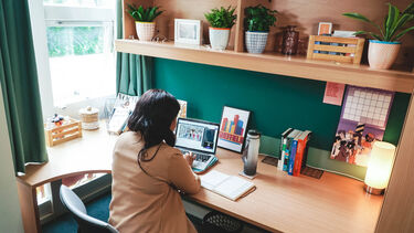 Young black girl sat at a desk working on a laptop next to a bright window. There are lots of plants on the shelves.