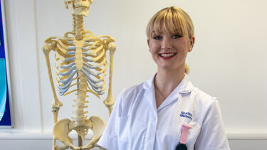 Isabel Gatenby has blonde hair tied back. She is wearing a white student midwife uniform and standing in front of an anatomical skeleton.