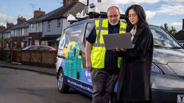 a man in a hi-vis vest looks over the shoulder of a woman, a researcher, who is holding a laptop. In the background is an electric van. 