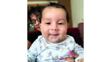 Baby Sebastian smiles at the camera in the foreground with his mum, Dharmisha, in the background