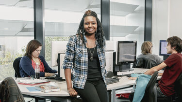 Staff in background sat around a desk, with one member of staff facing the camera, leaning against the desk