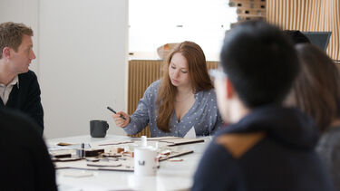 Shot of students at a table, looking over a small architecture model. 