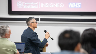 Male audience member with short grey hair wearing glasses and a dark suit, holds microphone to ask a question.