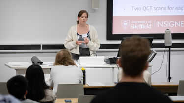 Female speaker  with brown hair tied in a ponytail wearing a light cardigan and striped top speaking to the audience in a lecture theatre