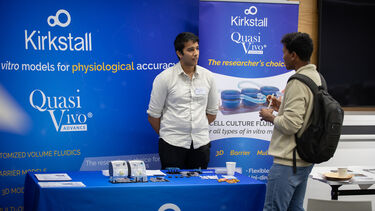 A man wiht dark hair wearing a white shirt stands in front of a blue company banner stand talking to an event attendess wearing a rucksack