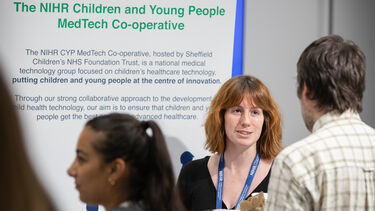 A woman with wavy red hear stands talking to a man with brown hair in front of a banner stand