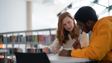 Two people sit at a desk with a bookshelf behind them. They seem to be working on something together.
