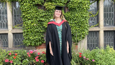 Young woman in university gown smiling in front of a uiversity building